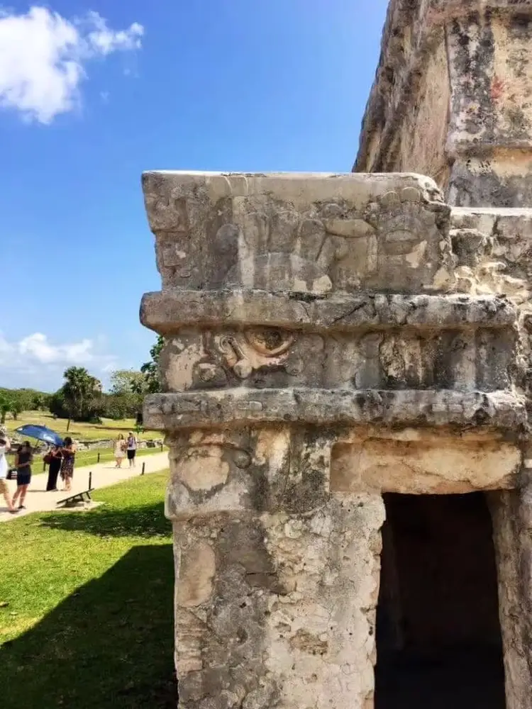Weathered stone column with carved details and a dark doorway opening, with visitors and green grounds visible in the background under blue sky Weathered stone column with carved details and a dark doorway opening, with visitors and green grounds visible in the background under blue sky
