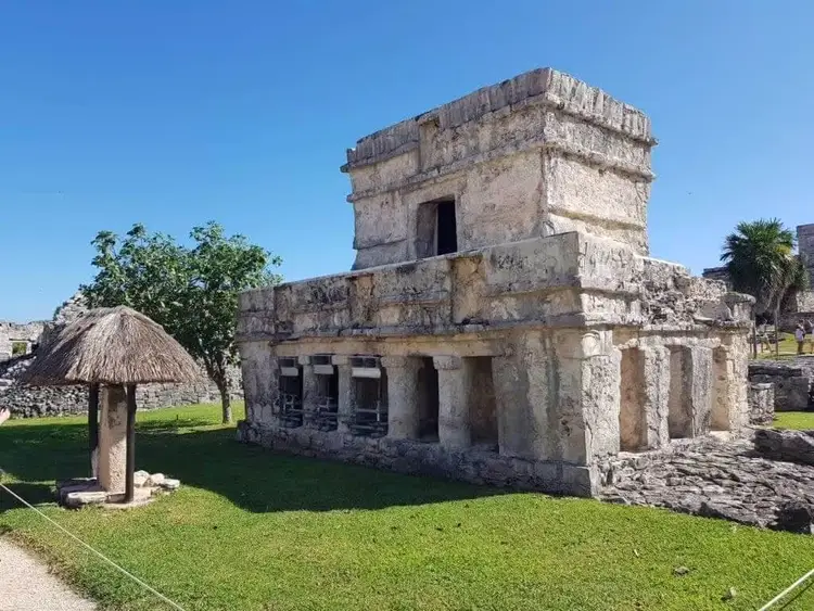 Ancient stone structure with tower stands on grassy grounds beside a thatched-roof building, under clear blue sky Ancient stone structure with tower stands on grassy grounds beside a thatched-roof building, under clear blue sky