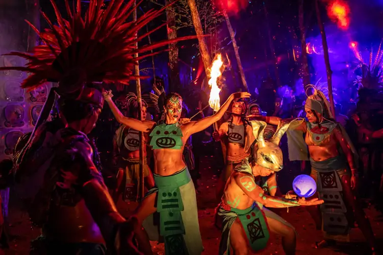 Performers in body paint and elaborate costumes dance with fire props at a nighttime festival stage surrounded by red and blue lighting