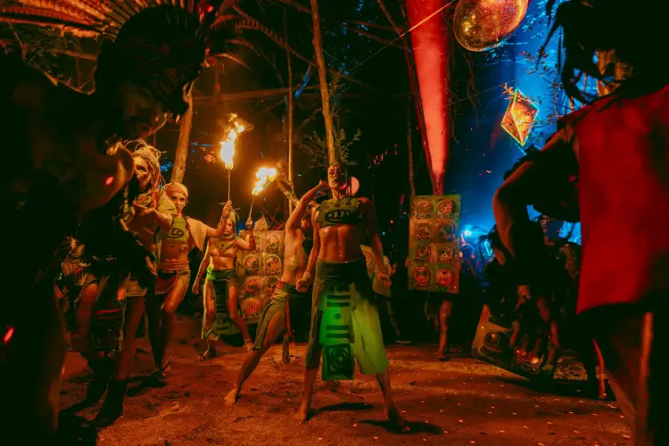 People in body paint and minimal clothing perform fire-spinning and dance at a nighttime outdoor festival event with colored lighting and decorations