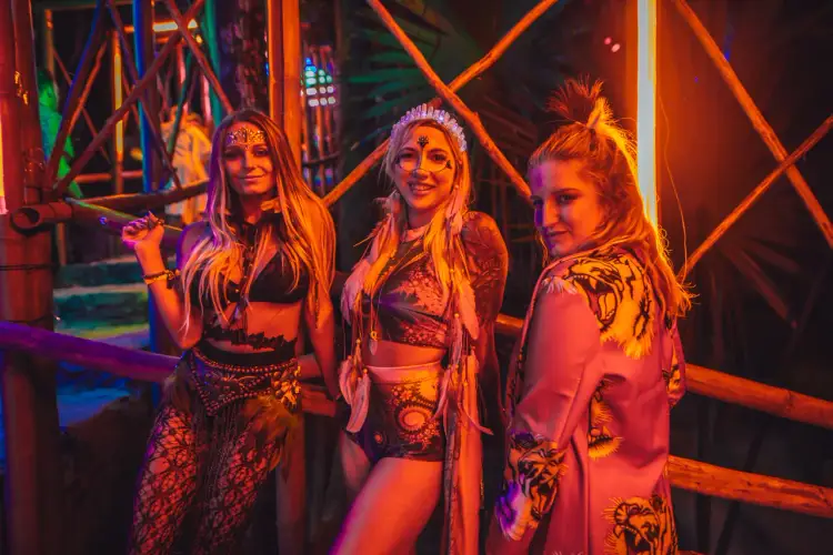 Three women pose together in a nightclub setting illuminated by orange and blue neon lights and geometric framework
