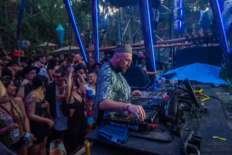 A DJ performs at an outdoor festival surrounded by crowds of attendees beneath blue lighting and trees