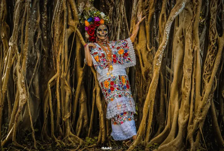 Person wearing skull face paint, floral dress and flower crown stands with arms outstretched among hanging dried vines
