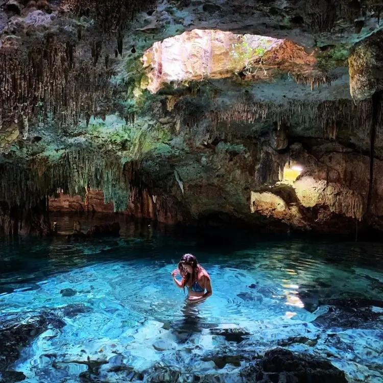 Person wading in clear turquoise water inside a cave with stalactites and an opening to daylight above