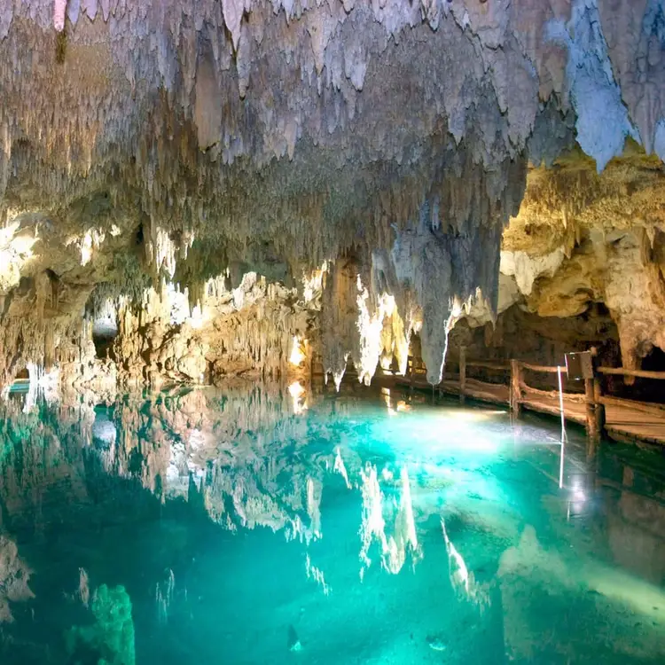 Cave interior with hanging stalactites and turquoise water below a wooden walkway along the cavern wall