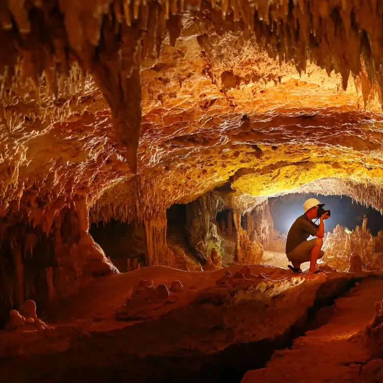 A person wearing a hard hat and headlamp crouches inside a cave chamber filled with stalactites and stalagmites illuminated in warm orange and yellow tones