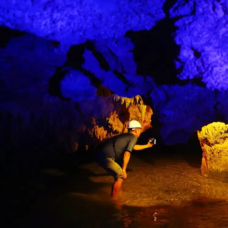 Person exploring underground cave chamber with blue-lit ceiling formations and golden-lit rock walls surrounding still water