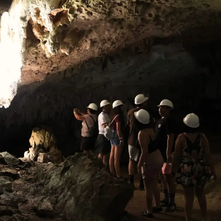 Group of visitors wearing hard hats standing inside a cave with limestone formations and natural light entering from an opening above