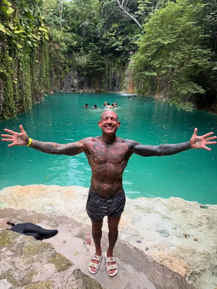 Man with outstretched arms stands on rocky shore of turquoise lagoon surrounded by dense forest vegetation, with other swimmers visible in the water behind him