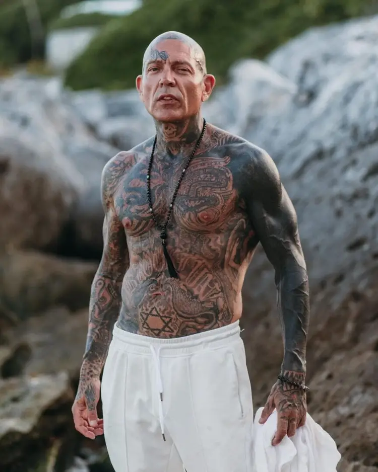 Shirtless man with extensive tattoos covering his torso and arms stands outdoors near large rocks with greenery in the background