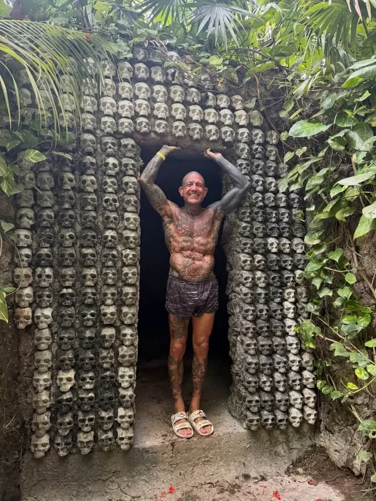 Man posing in doorway of stone structure decorated with carved skulls, surrounded by tropical vegetation