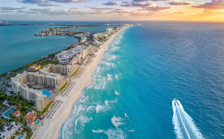 Aerial view of a beachfront resort area with high-rise hotels, sandy beach, turquoise water, and a speedboat creating a wake offshore at sunset Aerial view of a beachfront resort area with high-rise hotels, sandy beach, turquoise water, and a speedboat creating a wake offshore at sunset