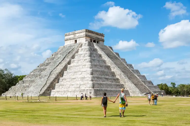 Visitors walk across a grassy plaza toward a large stepped pyramid with a temple structure on top under a blue sky Visitors walk across a grassy plaza toward a large stepped pyramid with a temple structure on top under a blue sky