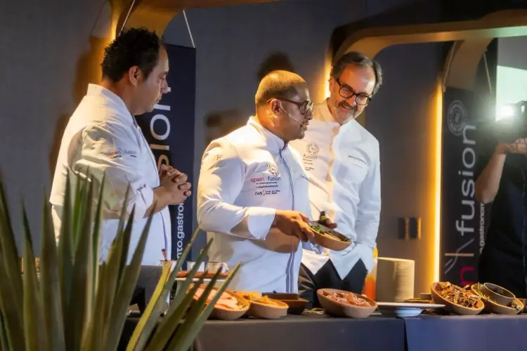 Three chefs in white chef's coats stand at a counter during what appears to be a culinary demonstration or event, with plated dishes visible on the table