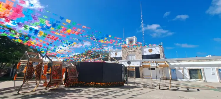 Colorful papel picado banners stretch across a rooftop plaza above traditional woven structures and modern architectural elements under a clear blue sky