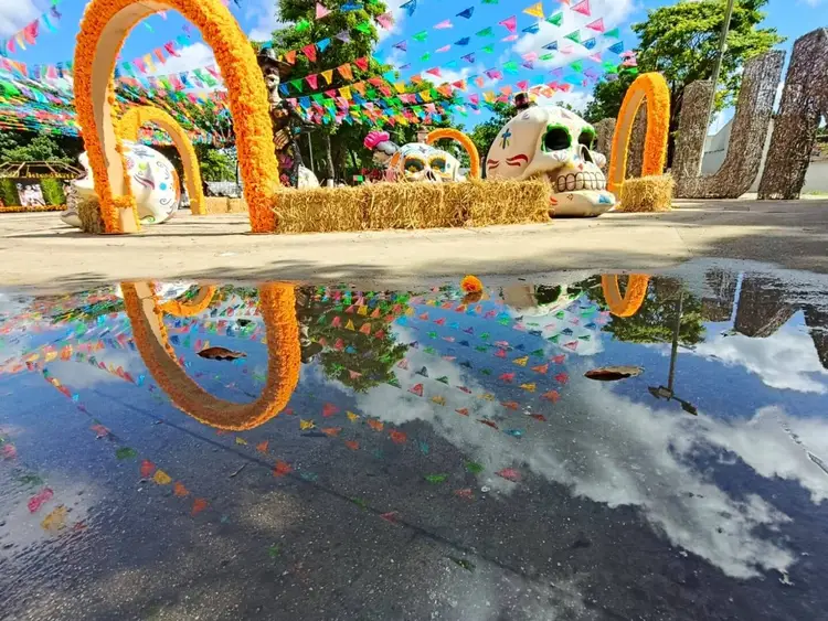 Outdoor installation with orange marigold arches, large skull decorations, colorful paper banners, and hay bales reflected in a puddle of water