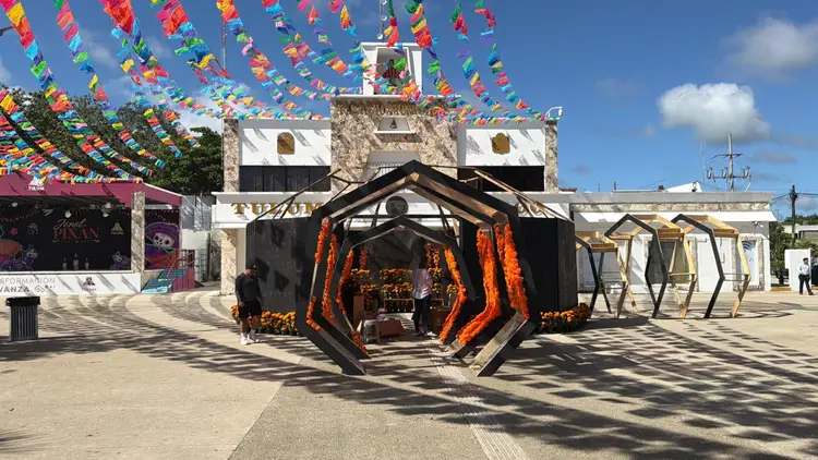 A geometric black sculptural structure decorated with orange marigolds stands in a plaza beneath colorful papel picado banners, with colonial architecture and w