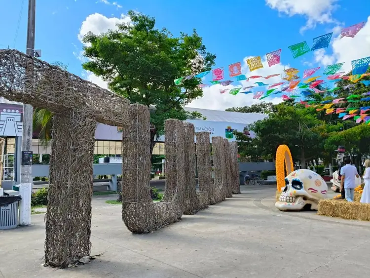 Outdoor plaza with tall woven cylindrical structures, colorful papel picado banners overhead, a decorated skull sculpture, and visitors walking through the spac