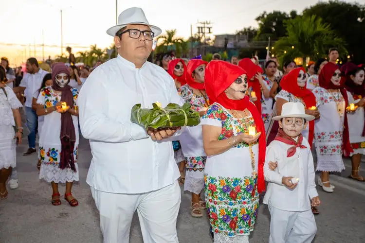 Man in white suit and hat holding vegetables walks with woman and child in colorful embroidered clothing and skull makeup during a Day of the Dead procession at