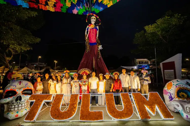 A group of people in light-colored clothing stand in front of a large wooden sign beneath an illuminated skeleton figure and colorful paper decorations at night