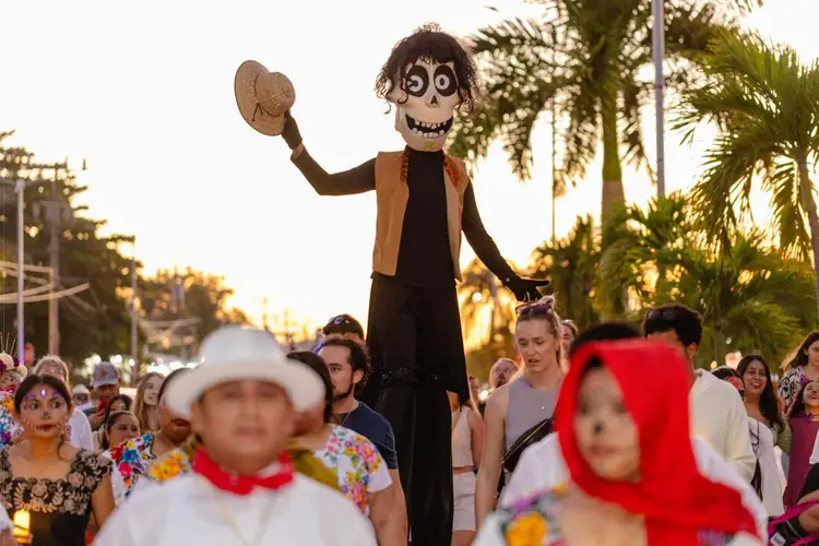 A skeletal puppet figure in a brown vest and black clothing holds a hat above a crowd of people wearing skull makeup and colorful clothing beneath palm trees at