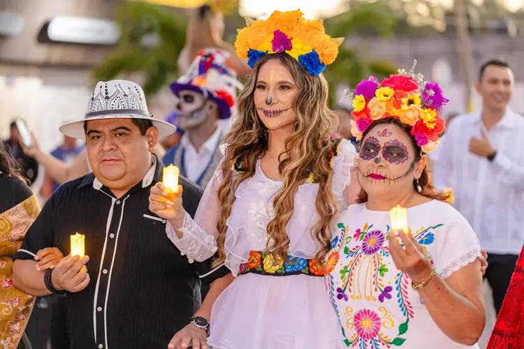 People wearing skull face paint and colorful flower crowns hold candles while participating in a Day of the Dead celebration outdoors