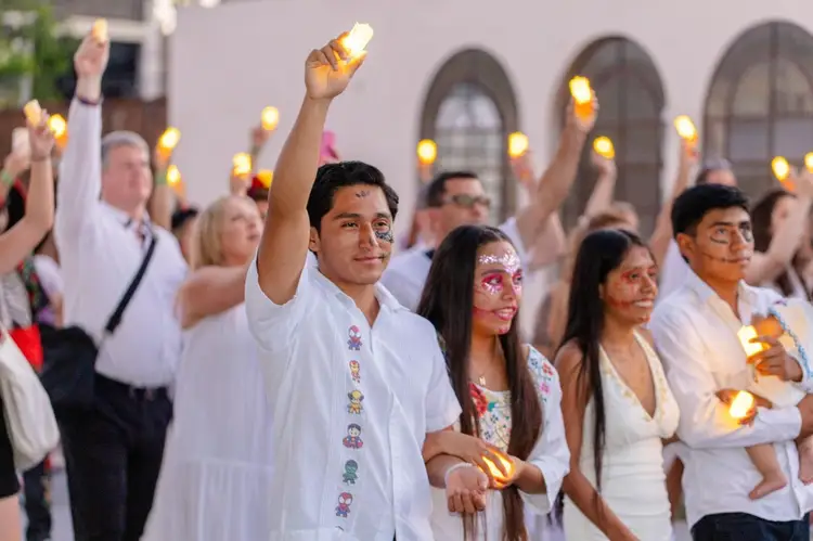 Crowd of people wearing white clothing and face paint hold lit candles while standing in front of a building with arched windows at dusk