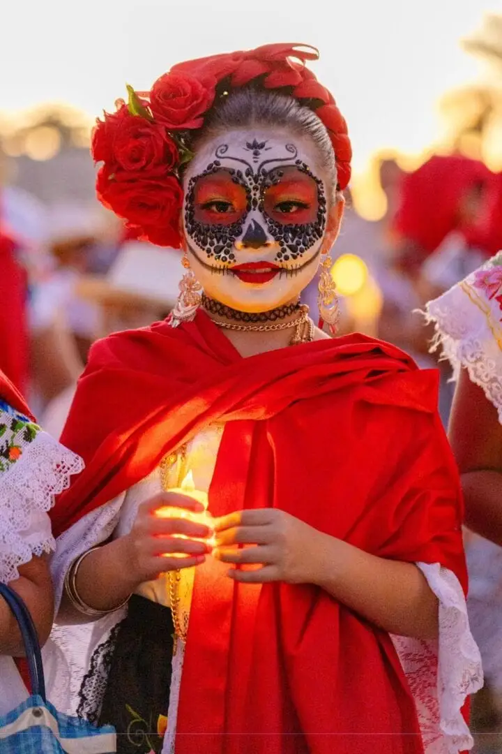 Person wearing skull face paint and red clothing with red roses in their hair holds a glowing candle at an outdoor gathering