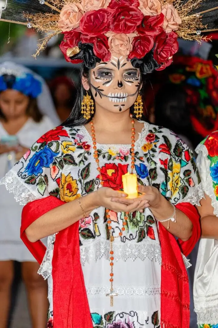 Person wearing skull face makeup and a large floral headdress holds a candle while dressed in an embroidered white blouse with red shawl and beaded necklace