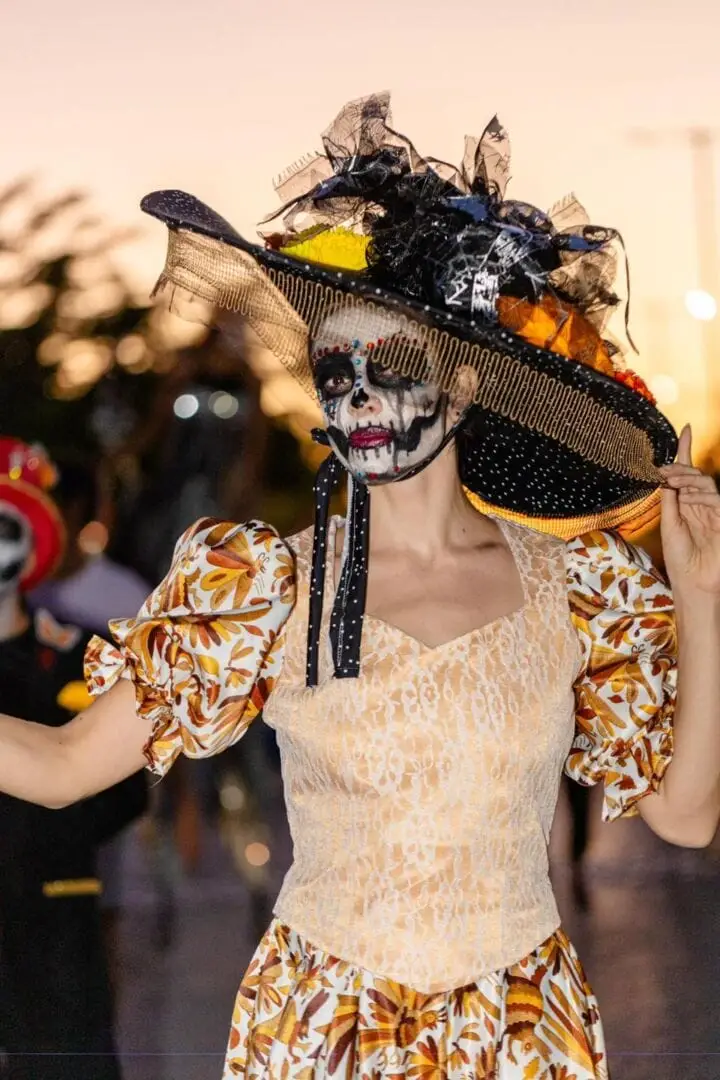 Person wearing skull face paint and an elaborate black headpiece adorned with colorful feathers and ornaments stands outdoors in costume