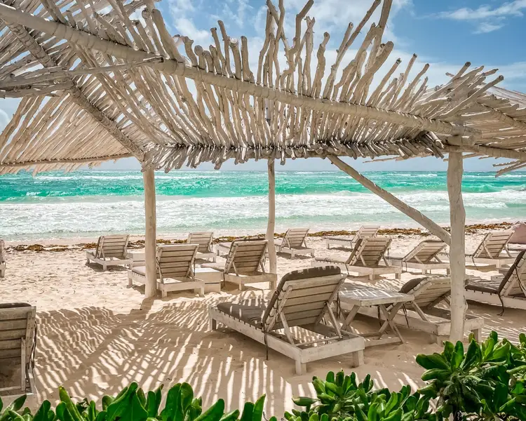 Wooden lounge chairs and shade structures line a sandy beach with turquoise water and whitecaps in the background Wooden lounge chairs and shade structures line a sandy beach with turquoise water and whitecaps in the background