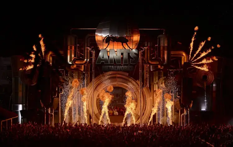 Large stage with ant logo and pyrotechnics firing flames during a nighttime concert performance with crowd in foreground Large stage with ant logo and pyrotechnics firing flames during a nighttime concert performance with crowd in foreground