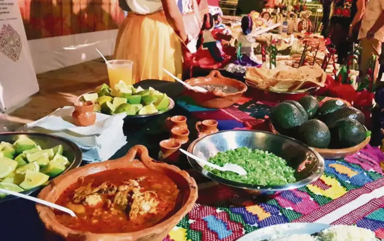 A spread of Mexican regional dishes displayed on colorful woven textiles, including stew in a clay bowl, fresh vegetables, and guacamole, with diners visible in