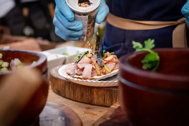 Chef in blue gloves plates a seafood dish on a wooden block surrounded by bowls of ingredients and garnishes