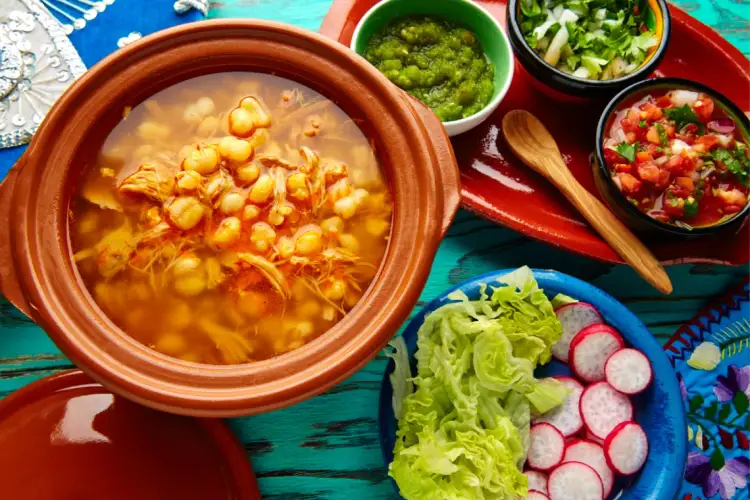 A terracotta bowl of chickpea stew sits on a turquoise wooden table surrounded by small bowls of green sauce, diced tomatoes, fresh lettuce and sliced radishes