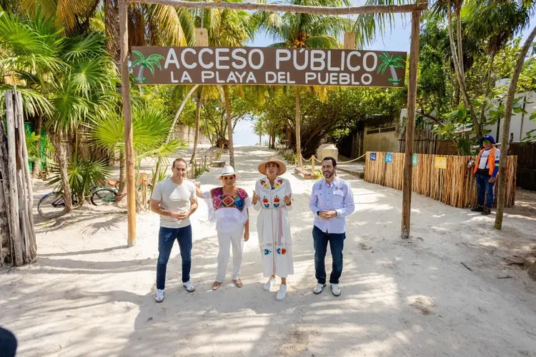Four people stand on a sandy path beneath a wooden archway with a sign reading "Acceso Público La Playa Del Pueblo," with palm trees and beach access visible be Four people stand on a sandy path beneath a wooden archway with a sign reading "Acceso Público La Playa Del Pueblo," with palm trees and beach access visible be