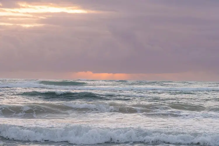 Waves roll toward shore under a cloudy sky with patches of golden light breaking through at dusk or dawn Waves roll toward shore under a cloudy sky with patches of golden light breaking through at dusk or dawn