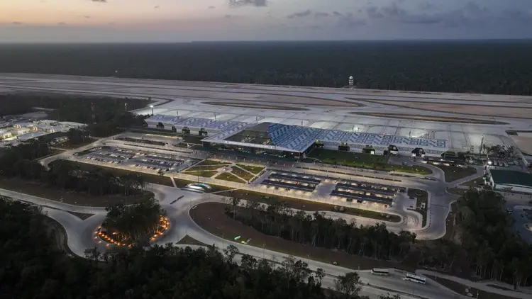 Aerial view of an airport facility at dusk with a racetrack-style road, large hangars, solar panels on roofing, and forested areas surrounding the complex