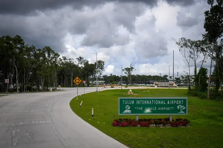 A green sign for Tulum International Airport sits on manicured grounds along a curved approach road, with airport facilities and planes visible in the distance
