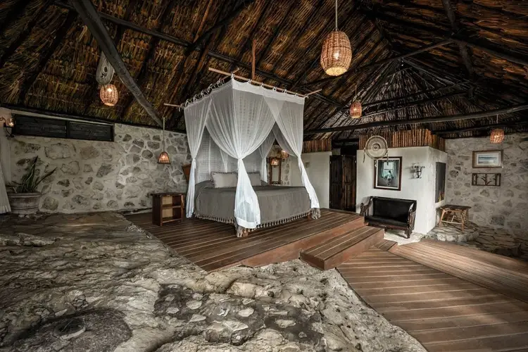 Interior of a hotel room with a four-poster bed draped in white fabric, woven pendant lights, and stone walls beneath a thatched roof Interior of a hotel room with a four-poster bed draped in white fabric, woven pendant lights, and stone walls beneath a thatched roof