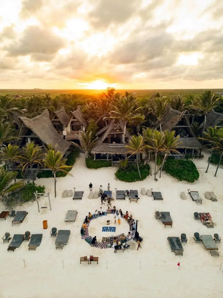 Aerial view of a beachfront resort with thatched-roof buildings, palm trees, and a group of people gathered in a circle on the sand at sunset Aerial view of a beachfront resort with thatched-roof buildings, palm trees, and a group of people gathered in a circle on the sand at sunset