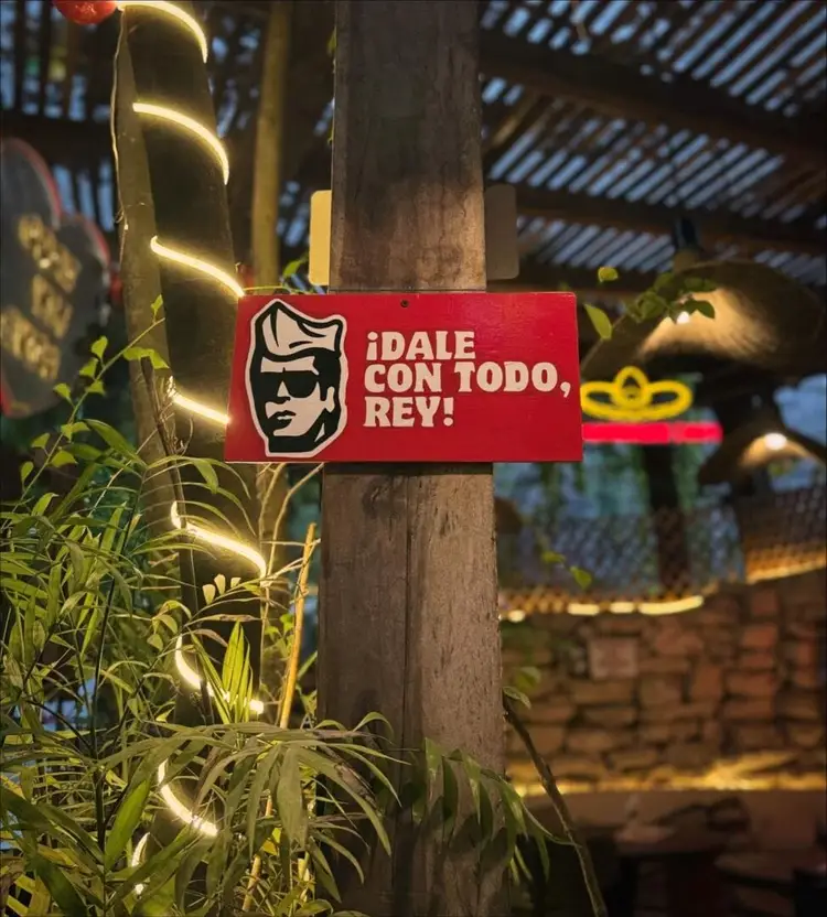 Red sign with Spanish text and an illustration attached to a wooden pole wrapped with glowing string lights, surrounded by green foliage in an outdoor venue set Red sign with Spanish text and an illustration attached to a wooden pole wrapped with glowing string lights, surrounded by green foliage in an outdoor venue set