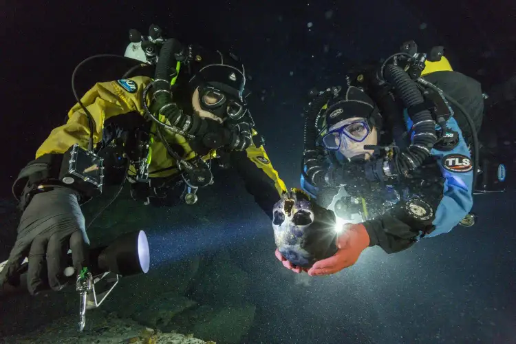Two scuba divers in full gear examine a skull underwater in a dark cave environment
