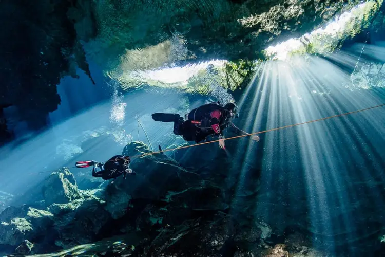 Two scuba divers explore an underwater cave system with sunlight streaming through openings above