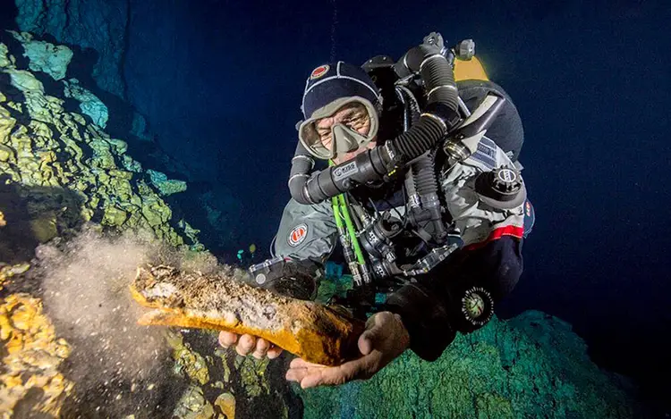 Scuba diver in full gear examines underwater rock formations covered in algae and mineral deposits in deep blue water