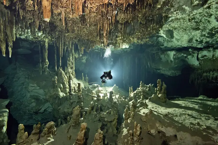 A diver explores an underwater cave system with stalactites hanging from the ceiling and rock formations rising from the cave floor