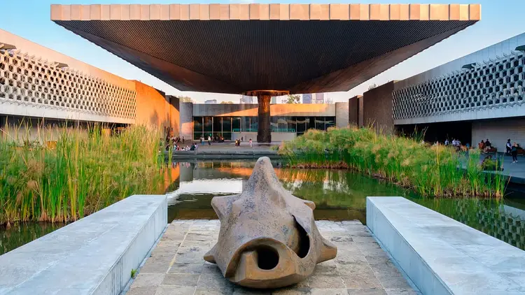 A stone sculpture sits on a platform between two white walls overlooking a pond with marsh grasses, with modern buildings and a large overhead structure visible A stone sculpture sits on a platform between two white walls overlooking a pond with marsh grasses, with modern buildings and a large overhead structure visible