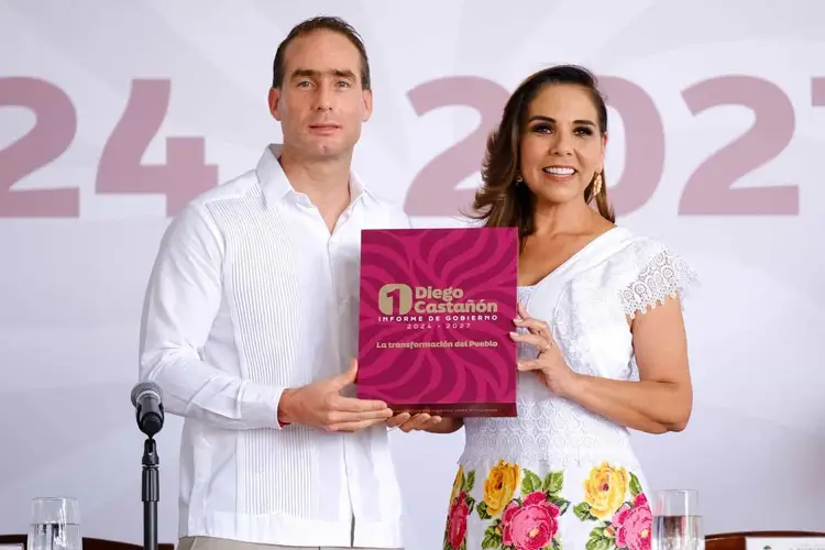 Two people in white clothing hold a burgundy folder at a formal event with numbered backdrop signage and microphone visible Two people in white clothing hold a burgundy folder at a formal event with numbered backdrop signage and microphone visible
