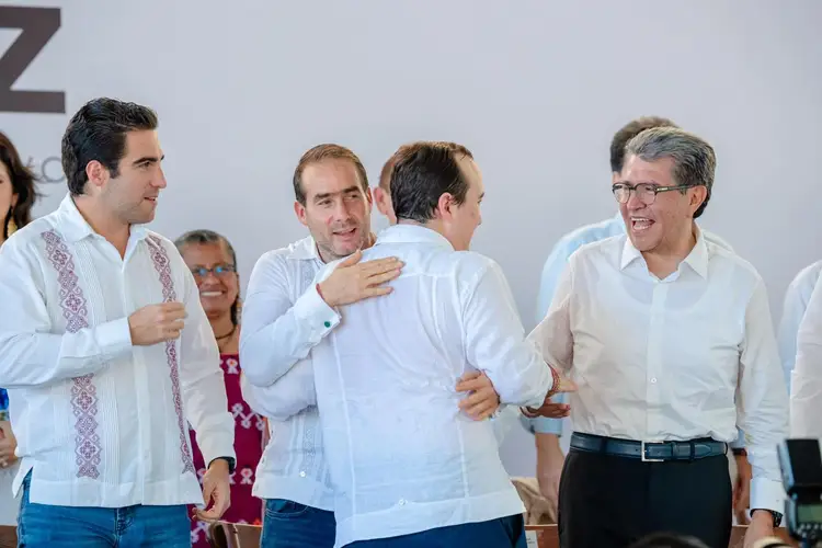 Four men in white shirts stand together at a public event, with one man in the center embracing another while others look on
