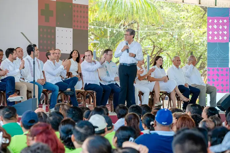 Man in light blue shirt and dark pants speaks at microphone to seated panel and crowd of onlookers at outdoor event under trees